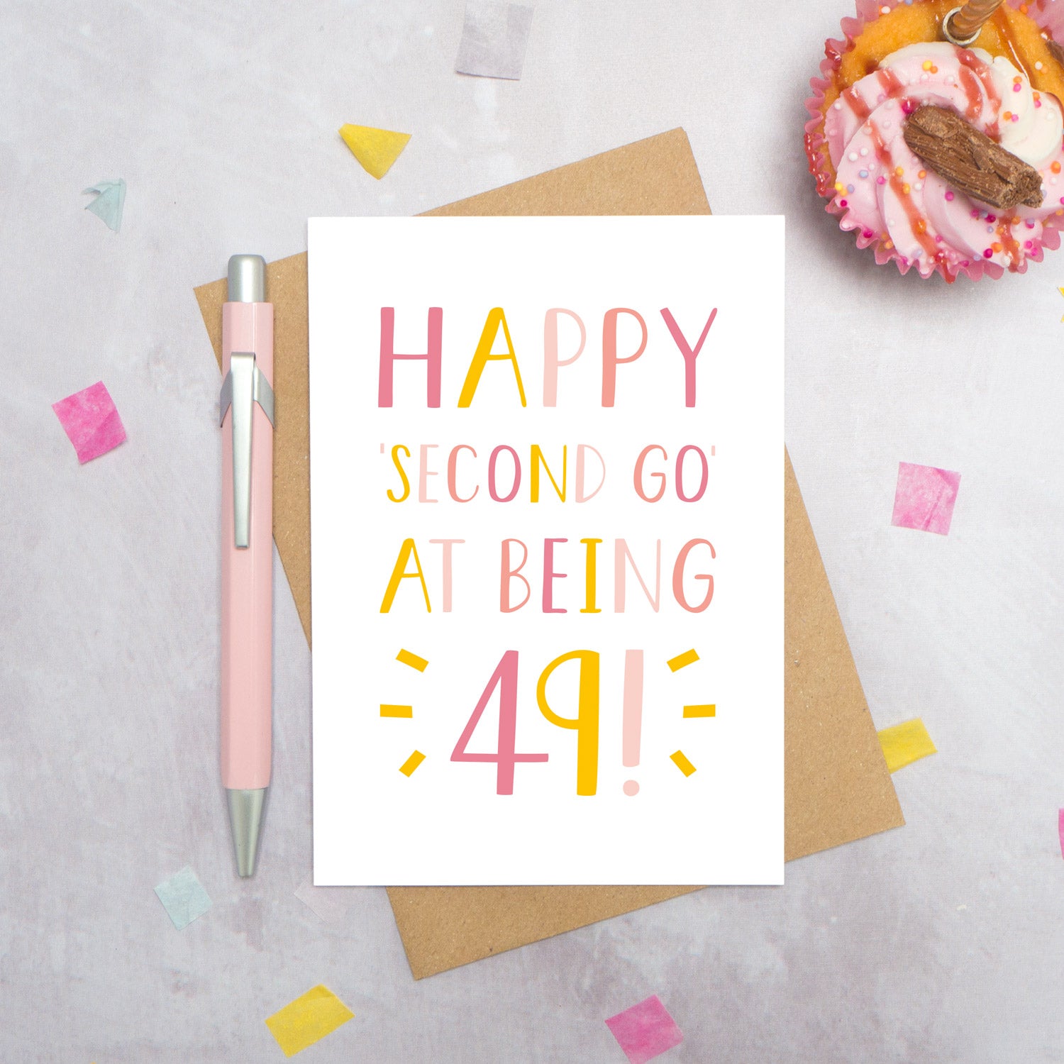 Happy second go at being 49 - milestone age card in pink photographed on a grey background surrounded by a cupcake, pen and confetti.