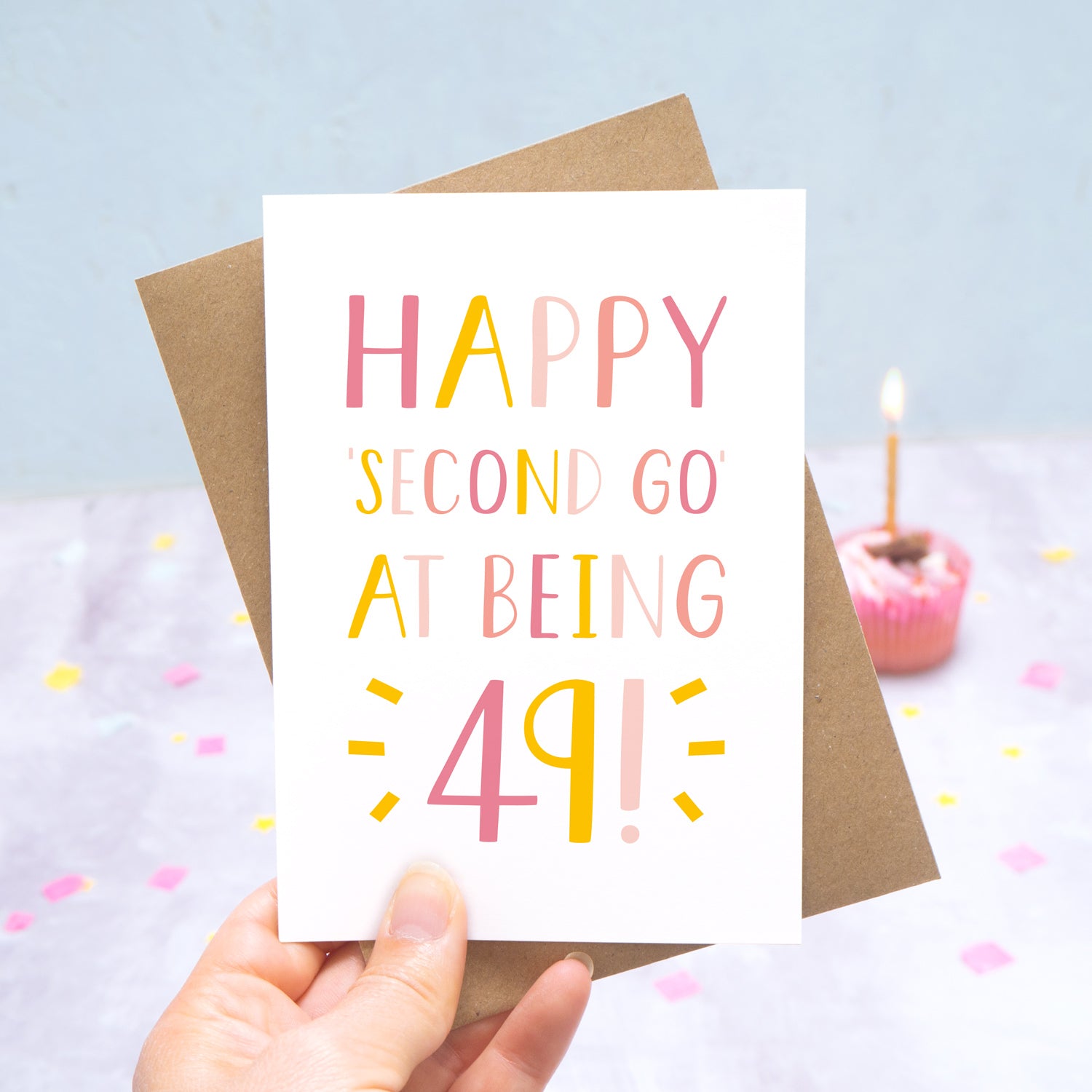 Happy second go at being 49 - milestone age card in pink photographed on a grey and blue background with a cupcake and burning candle.