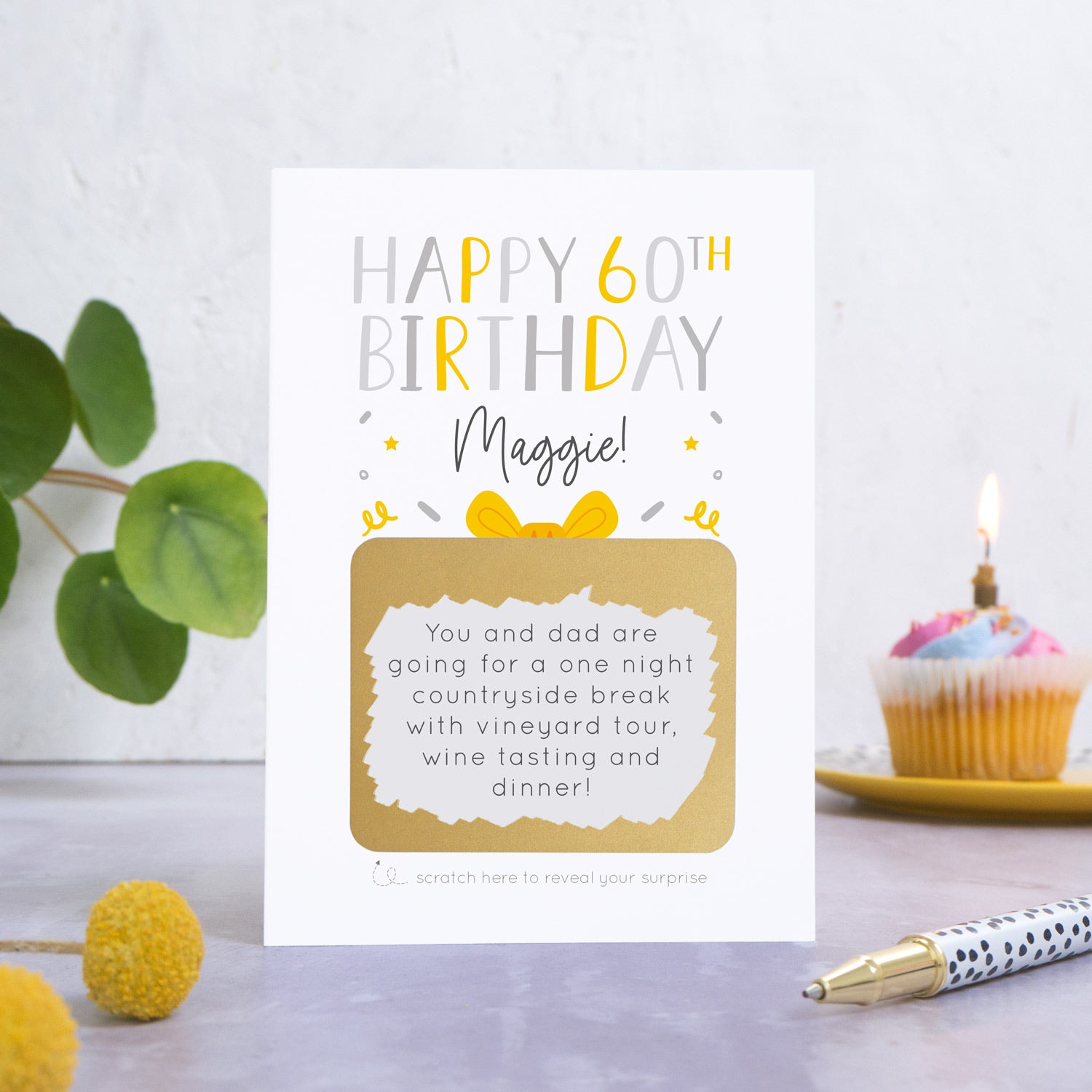 A personalised happy 60th birthday scratch card in grey that has been photographed on a white and grey background. There is foliage and yellow flowers on the left and a cupcake and a pen to the right. The card features the recipients name and a scratch panel that has been scratched off revealing a personalised message.
