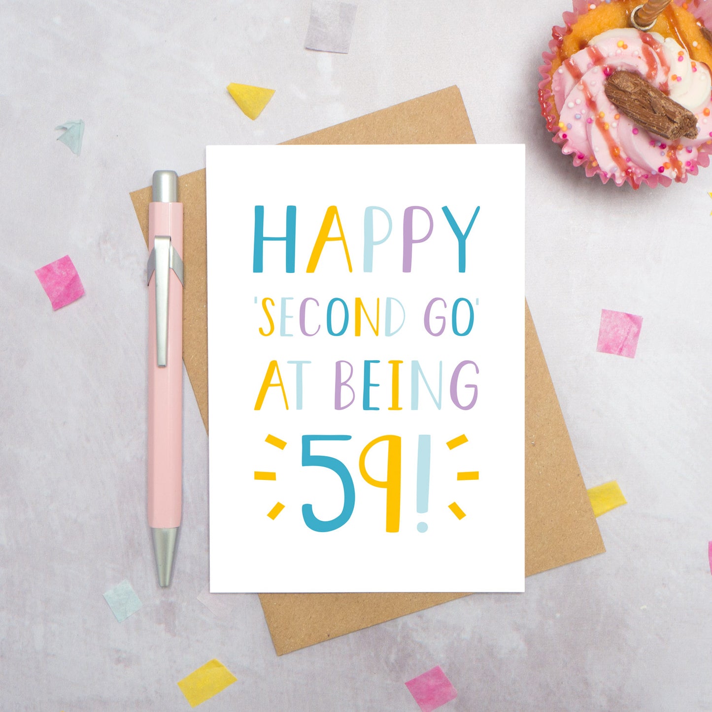 Happy second go at being 59 - milestone age card in blue, yellow and purple photographed on a grey background surrounded by a cupcake, pen and confetti.