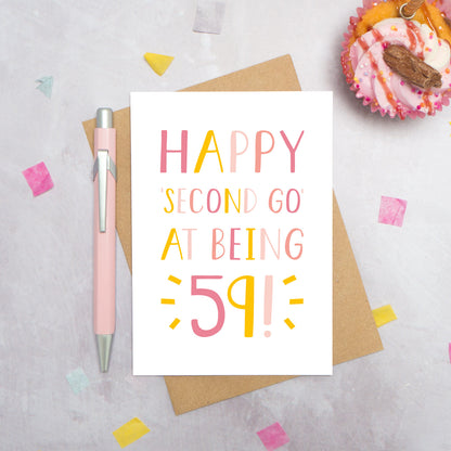 Happy second go at being 59 - milestone age card in pink photographed on a grey background surrounded by a cupcake, pen and confetti.