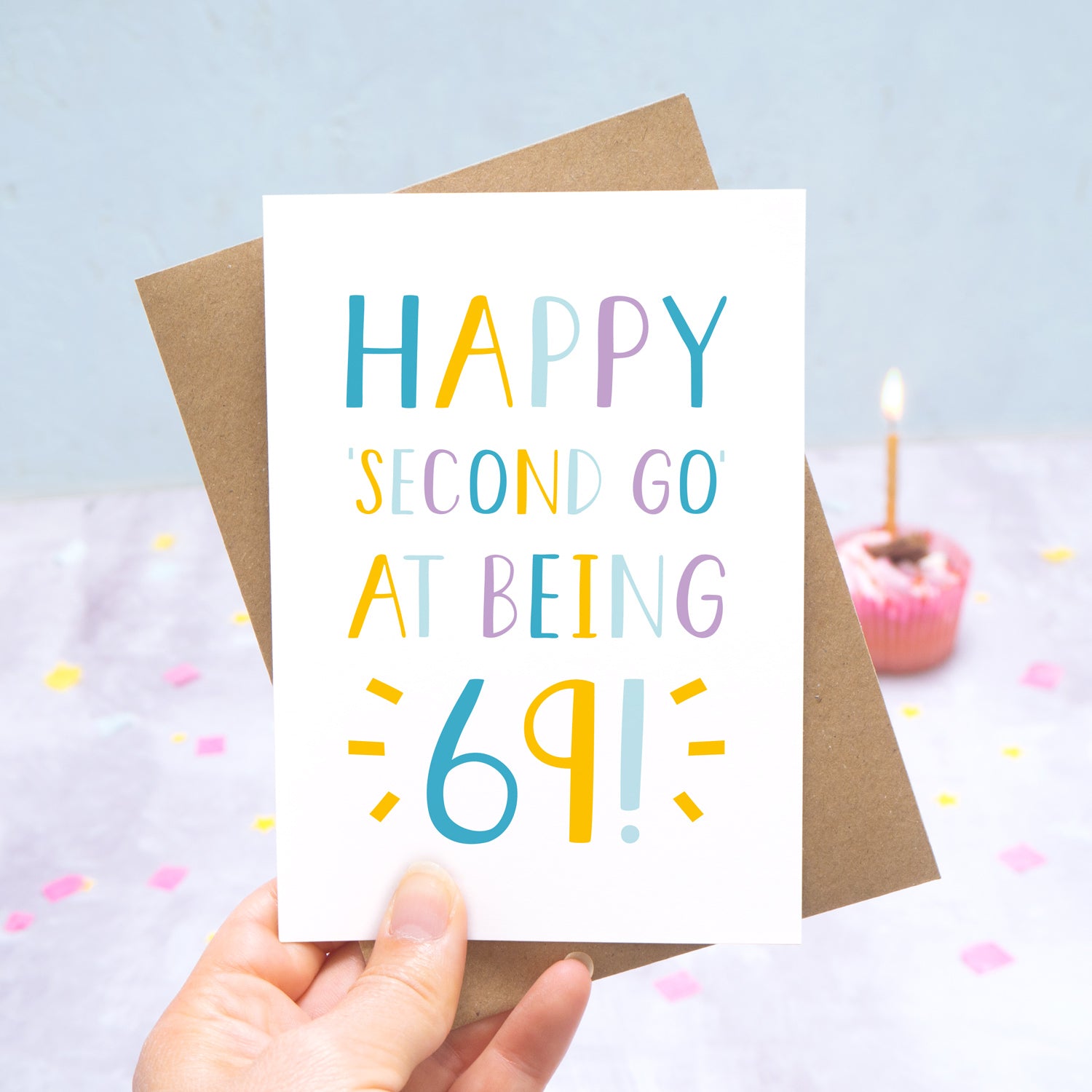 Happy second go at being 69 - milestone age card in blue, yellow and purple photographed on a grey and blue background with a cupcake and burning candle.