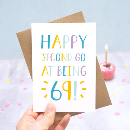 Happy second go at being 69 - milestone age card in blue, yellow and purple photographed on a grey and blue background with a cupcake and burning candle.