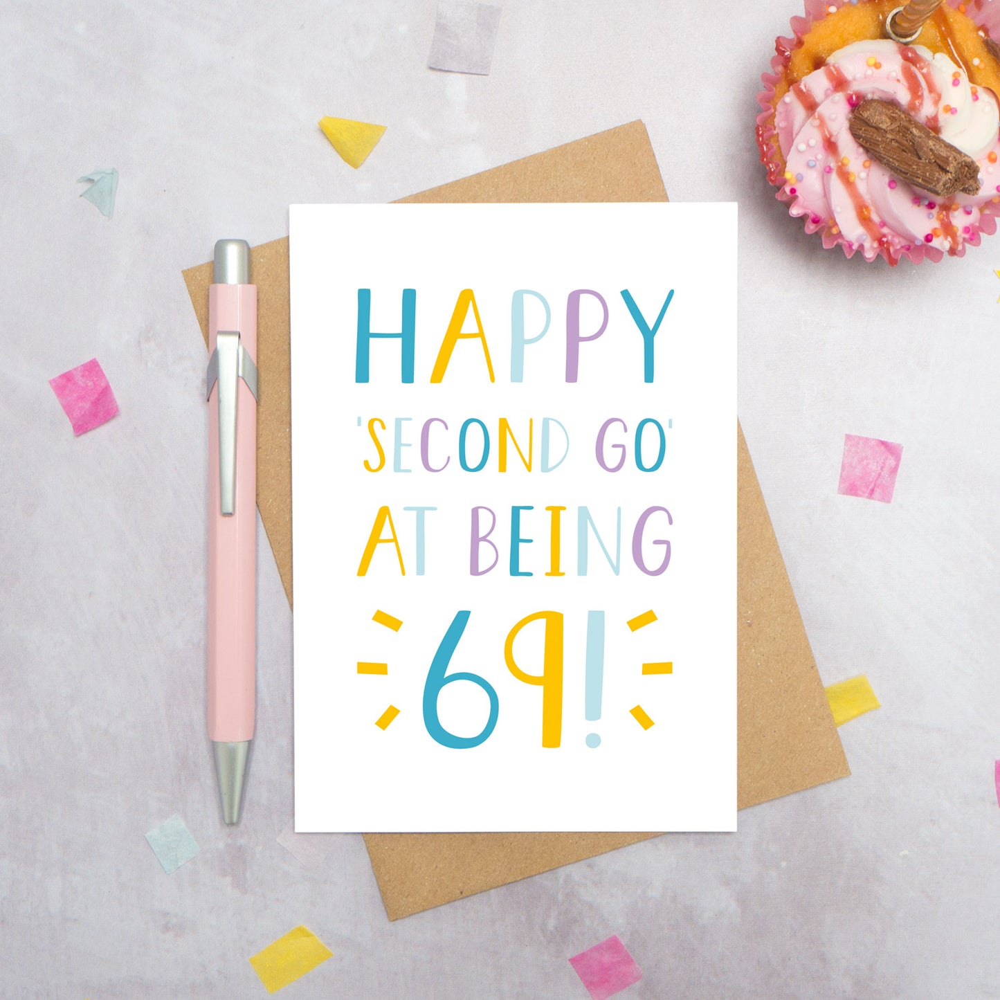 Happy second go at being 69 - milestone age card in blue, yellow and purple photographed on a grey background surrounded by a cupcake, pen and confetti.