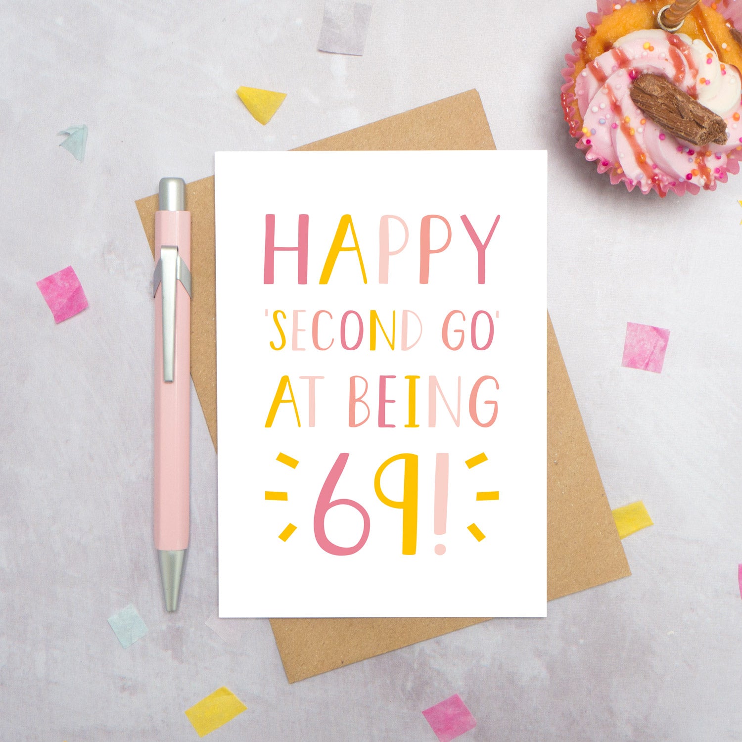 Happy second go at being 69 - milestone age card in pink photographed on a grey background surrounded by a cupcake, pen and confetti.