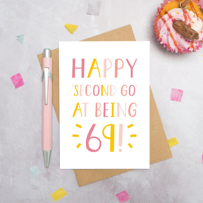 Happy second go at being 69 - milestone age card in pink photographed on a grey background surrounded by a cupcake, pen and confetti.