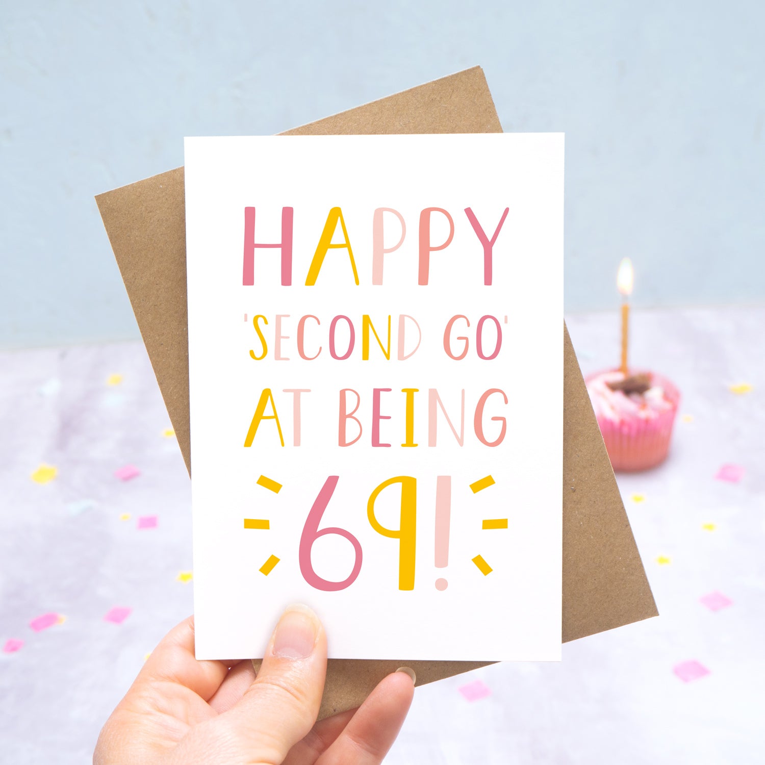 Happy second go at being 69 - milestone age card in pink photographed on a grey and blue background with a cupcake and burning candle.