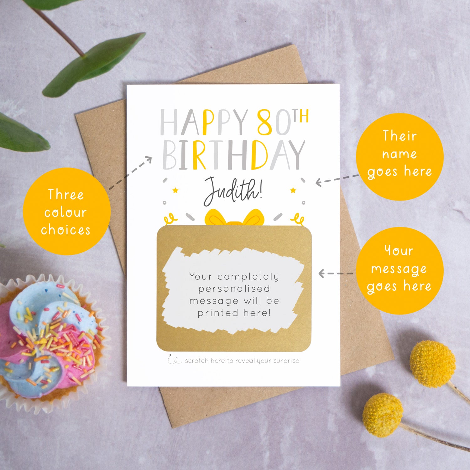 A personalised happy 80th birthday scratch card in grey that has been photographed on a grey background with foliage and a cupcake. There are also orange circles laid over the top of this image with arrows pointing to the areas that can be customised. In this instance they point to the name, the colour choices and the personalised scratch off message.
