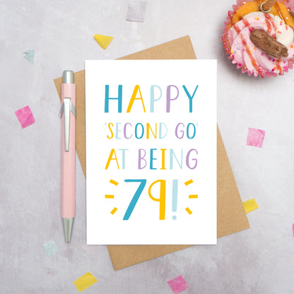 Happy second go at being 79 - milestone age card in blue, yellow and purple photographed on a grey background surrounded by a cupcake, pen and confetti.