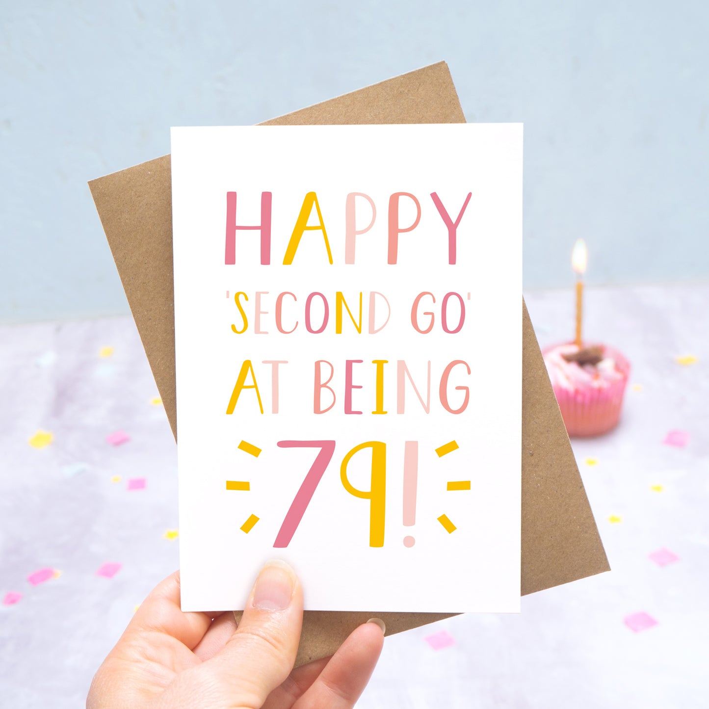 Happy second go at being 79 - milestone age card in pink photographed on a grey and blue background with a cupcake and burning candle.