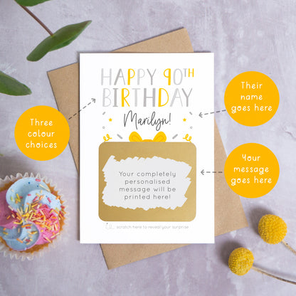 A personalised happy 90th birthday scratch card in grey that has been photographed on a grey background with foliage and a cupcake. There are also orange circles laid over the top of this image with arrows pointing to the areas that can be customised. In this instance they point to the name, the colour choices and the personalised scratch off message.