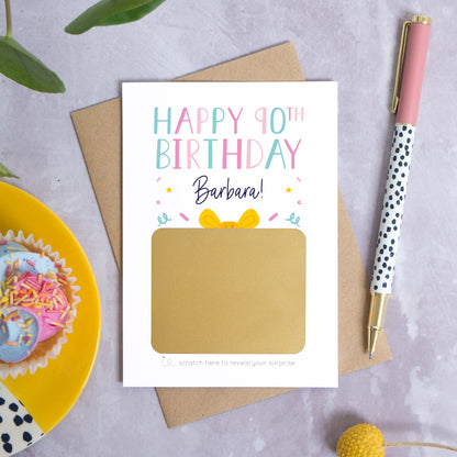 A personalised happy 90th birthday scratch card that has been photographed flat lay style on a grey concrete style background surrounded with foliage, a cupcake and a pen. The card itself shows how it will arrive and looks when it is completely unscratched.