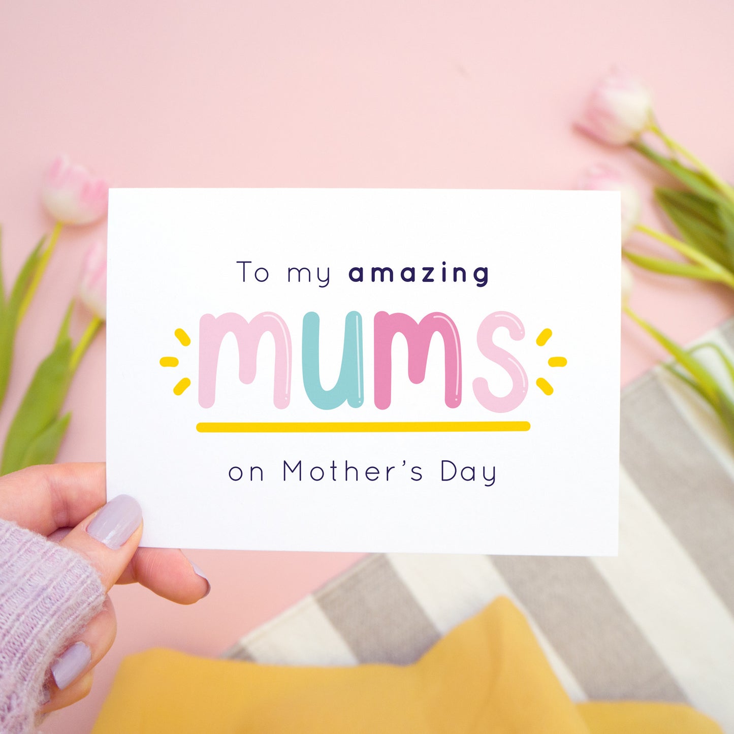 An amazing mums mother’s day card designed for those lucky enough to have two mums! This card is being held over a pink background with a grey and white rug with tulips to the side. This card is in the pink and blue colour palette.