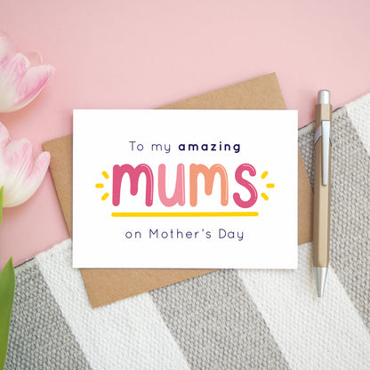 An amazing mums mother’s day card designed for those lucky enough to have two mums! This card is lying flat on top of its kraft brown envelope on top of a pink background with a grey and white rug with tulips to the left and a pen to the right. This card is in the pink and peach colour palette.