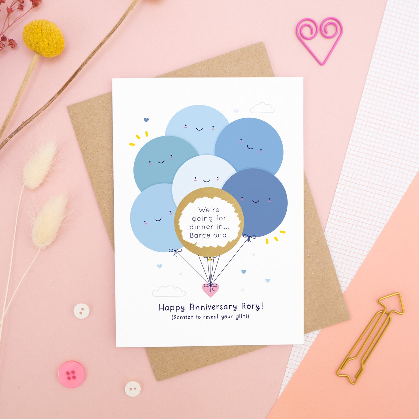 A personalised anniversary scratch card photographed on a pink background with floral props, paper clips, and buttons. This card shows the blue colour scheme and the golden circle has been scratched off to reveal the secret message!