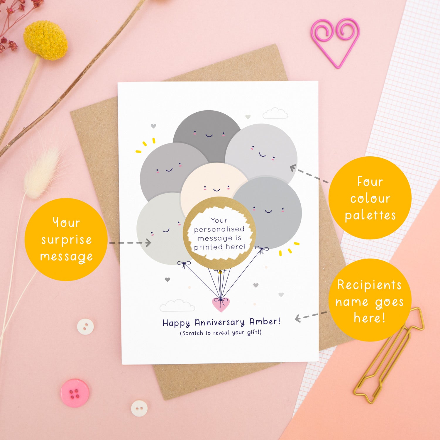A personalised anniversary scratch card photographed on a pink background with floral props, paper clips, and buttons. This card shows the grey colour scheme and the golden circle has been scratched off to reveal the secret message! Around the card are circles of text pointing to the areas that can be changed (name, colour and personalised message).