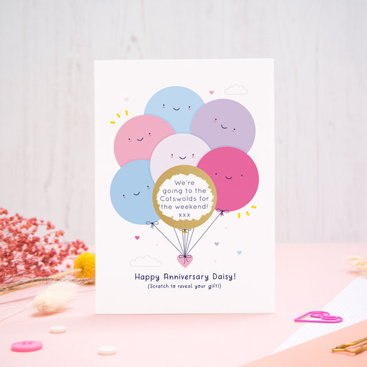 A personalised anniversary scratch card photographed on a pink and white background with floral props, paper clips, and buttons. This card shows the pink & lilac colour scheme and the golden circle has been scratched off to reveal the secret message!