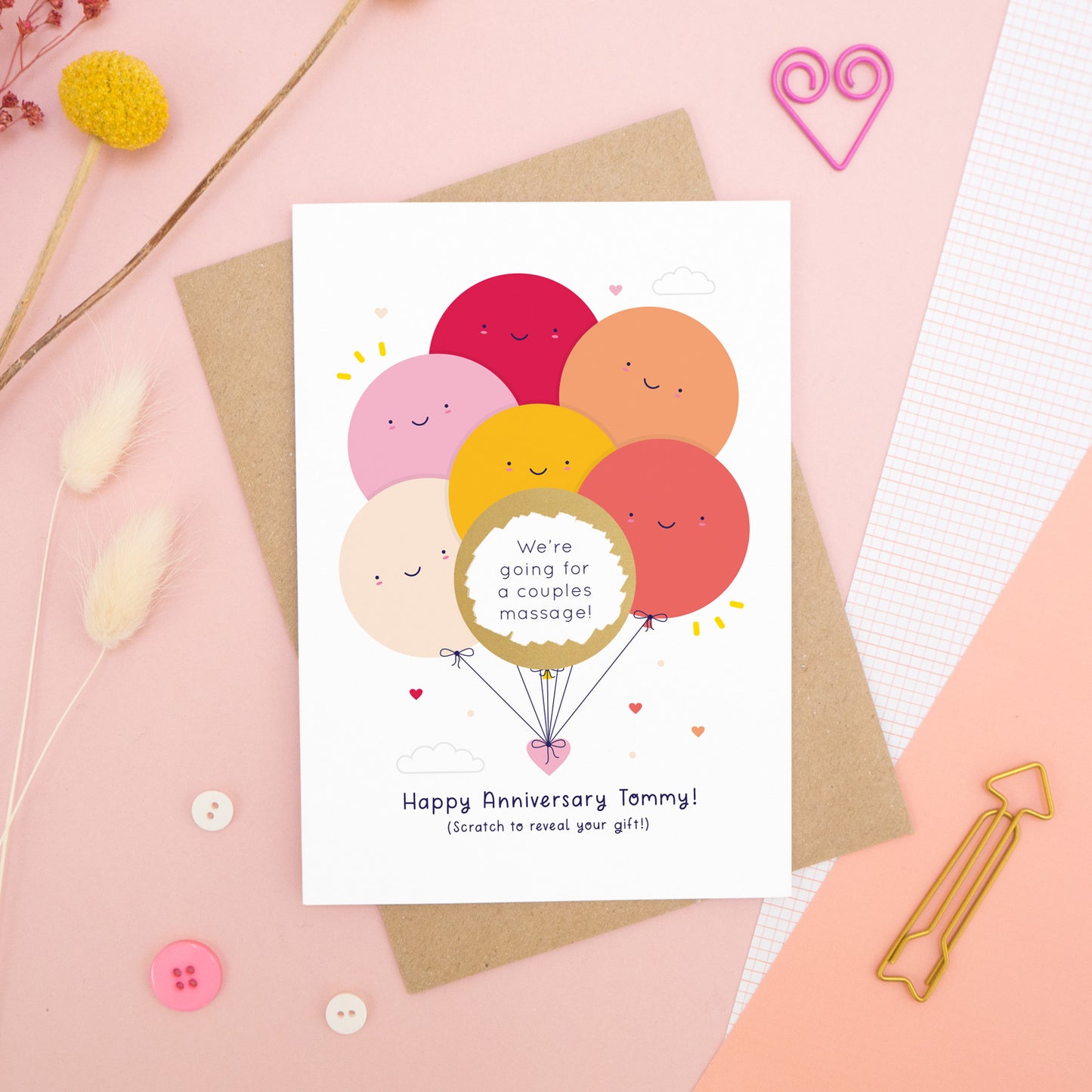 A personalised anniversary scratch card photographed on a pink background with floral props, paper clips, and buttons. This card shows the red & peach colour scheme and the golden circle has been scratched off to reveal the secret message!