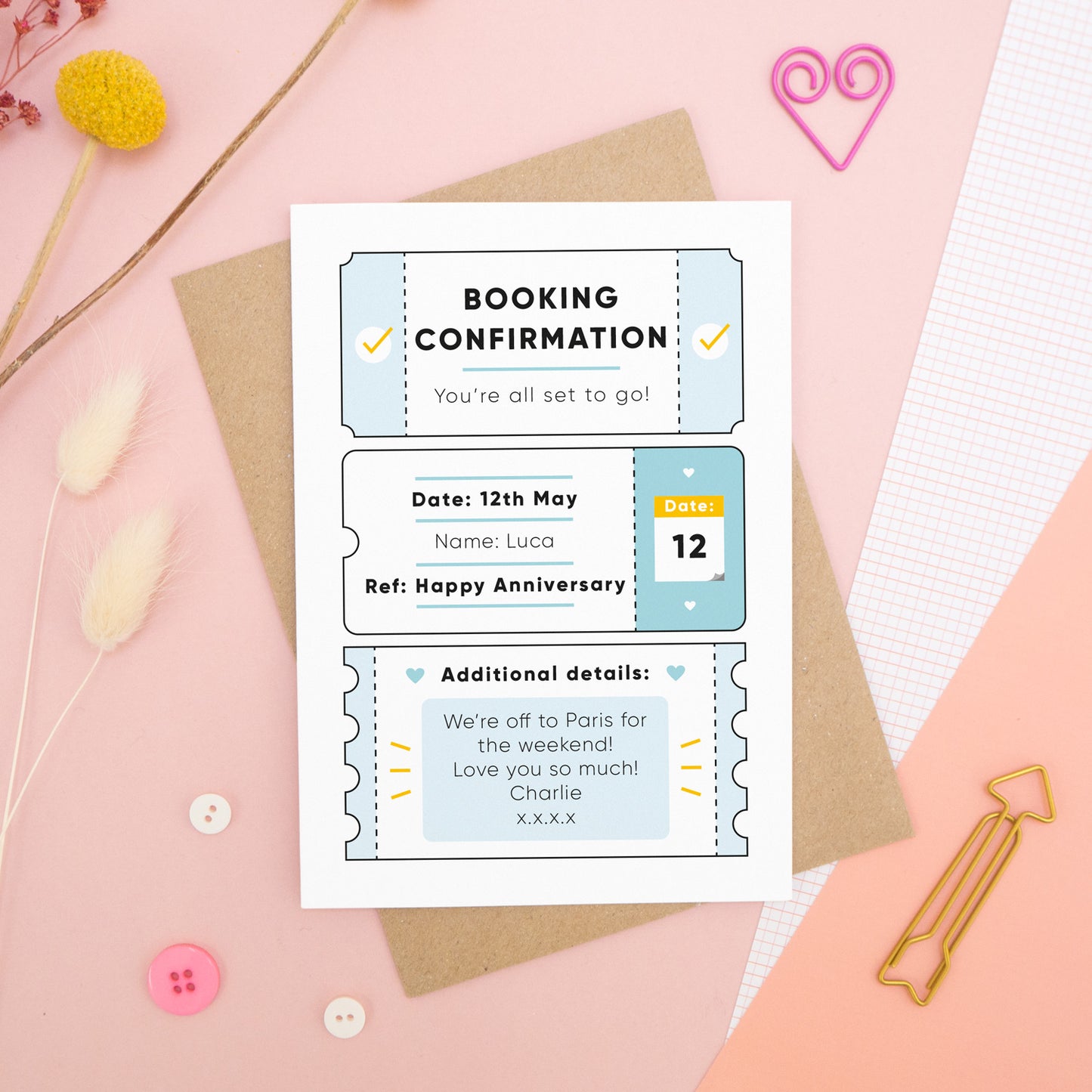 A personalised anniversary booking confirmation gift card lying flat lay on a pink and peach background scattered with dry flowers, grid paper, buttons and a heart. The card is lying on it’s kraft brown envelope and pictured is the blue version of the gift card.