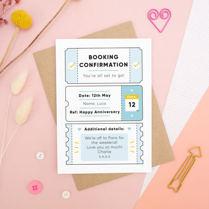A personalised anniversary booking confirmation gift card lying flat lay on a pink and peach background scattered with dry flowers, grid paper, buttons and a heart. The card is lying on it’s kraft brown envelope and pictured is the blue version of the gift card.