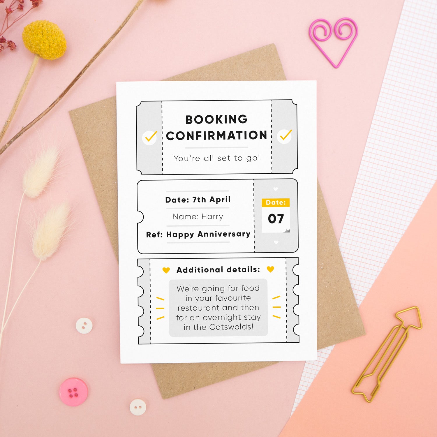 A personalised anniversary booking confirmation gift card lying flat lay on a pink and peach background scattered with dry flowers, grid paper, buttons and a heart. The card is lying on it’s kraft brown envelope and pictured is the grey version of the gift card.