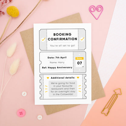 A personalised anniversary booking confirmation gift card lying flat lay on a pink and peach background scattered with dry flowers, grid paper, buttons and a heart. The card is lying on it’s kraft brown envelope and pictured is the grey version of the gift card.