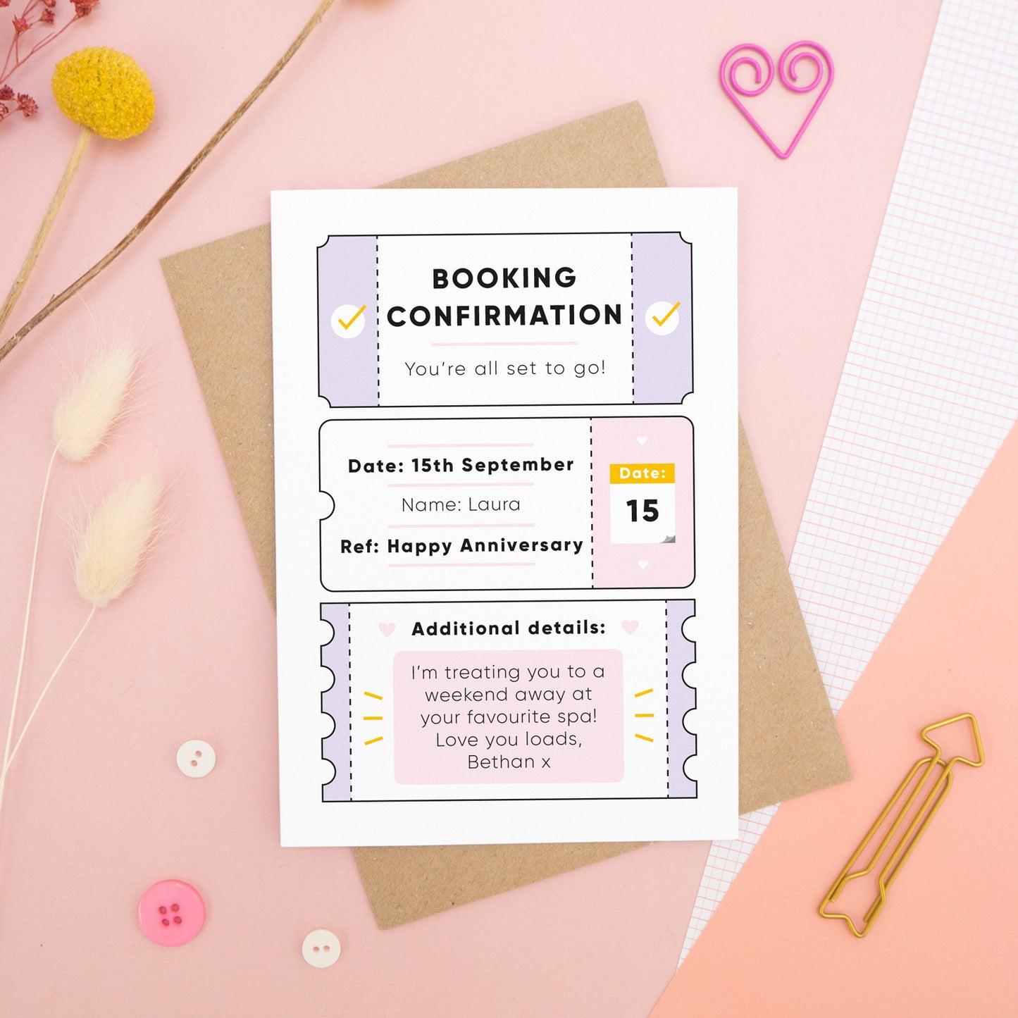 A personalised anniversary booking confirmation gift card lying flat lay on a pink and peach background scattered with dry flowers, grid paper, buttons and a heart. The card is lying on it’s kraft brown envelope and pictured is the lilac version of the gift card.