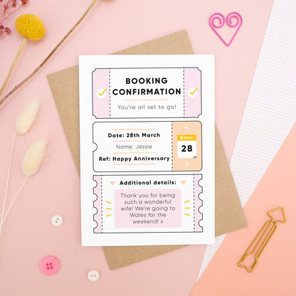 A personalised anniversary booking confirmation gift card lying flat lay on a pink and peach background scattered with dry flowers, grid paper, buttons and a heart. The card is lying on it’s kraft brown envelope and pictured is the pink version of the gift card.