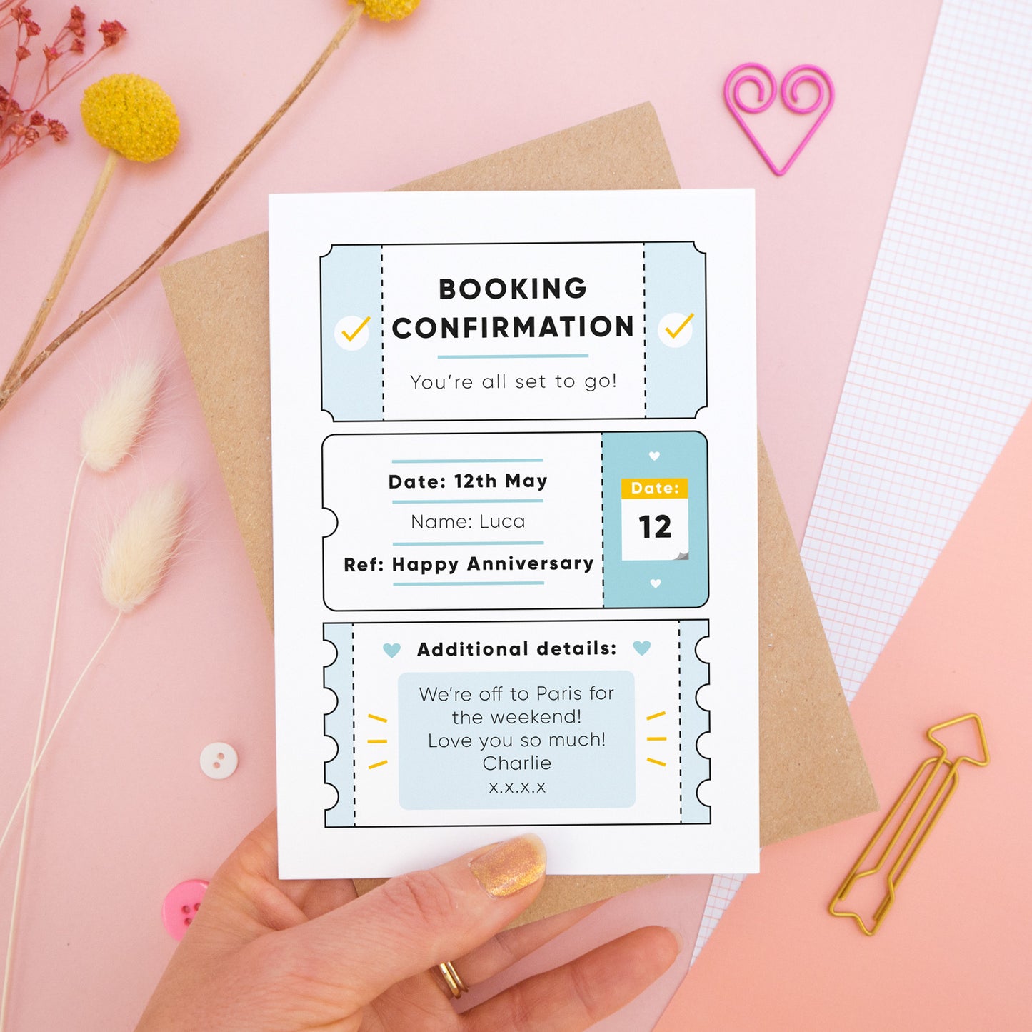 A personalised anniversary booking confirmation gift card held in front of a pink background scattered with dry flowers, grid paper, buttons and a heart. Pictured is the blue version of the card on top of a kraft brown envelope.
