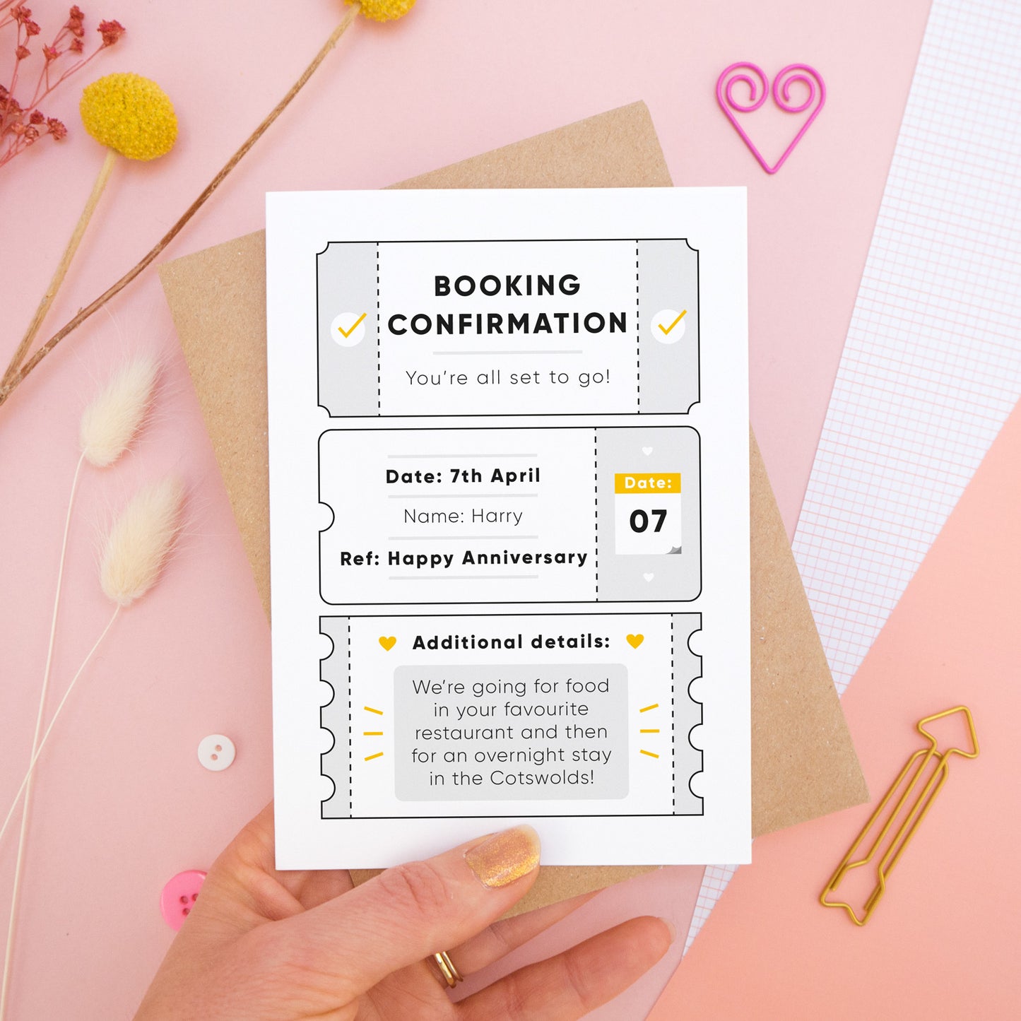 A personalised anniversary booking confirmation gift card held in front of a pink background scattered with dry flowers, grid paper, buttons and a heart. Pictured is the grey version of the card on top of a kraft brown envelope.