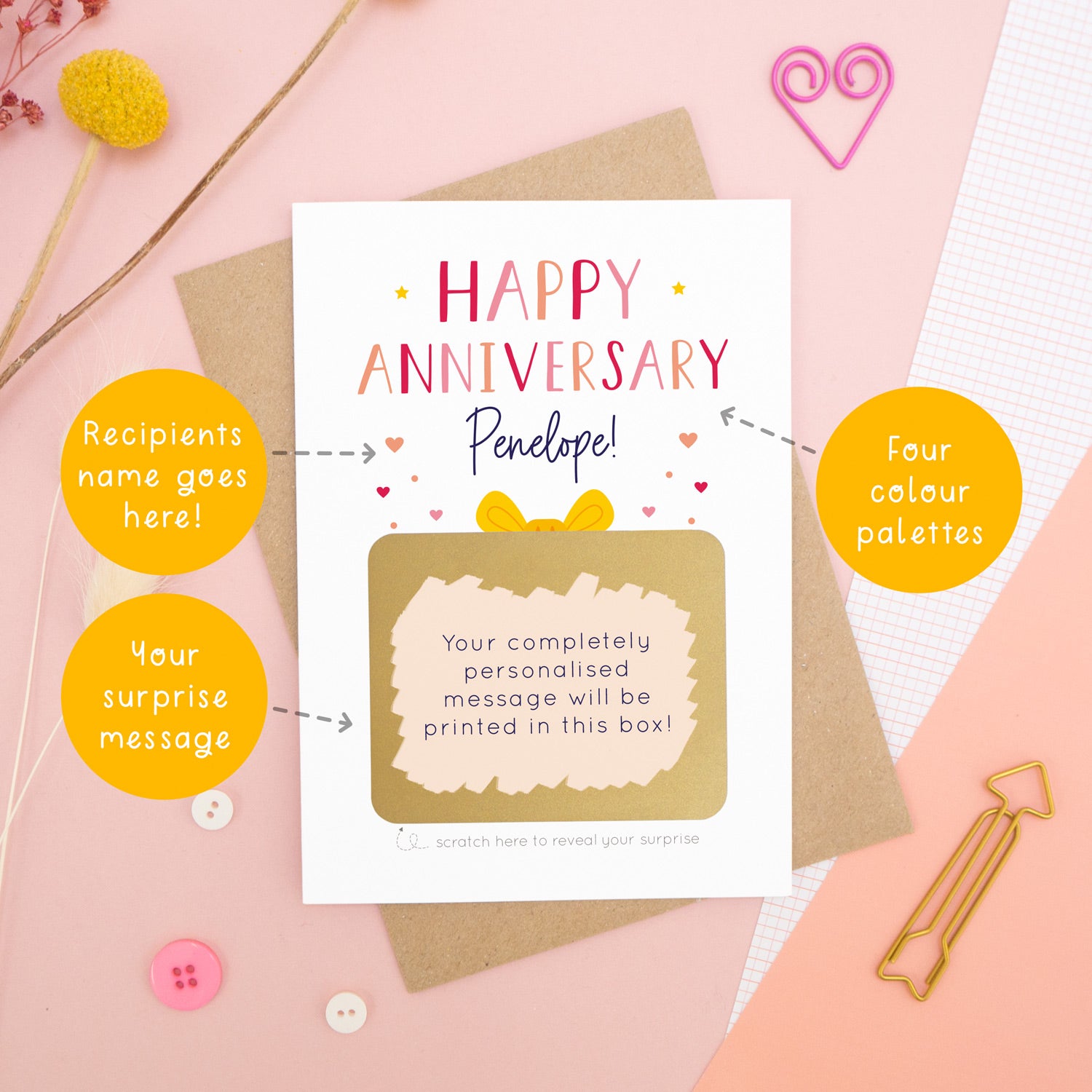 A personalised happy anniversary scratch card photographed on a pink background with floral props, paper clips, and buttons. The card has been scratched off to reveal the hidden message and is in the red and peach colour scheme. Around the card are circles of text pointing to the areas that can be changed (name, colour and personalised message).