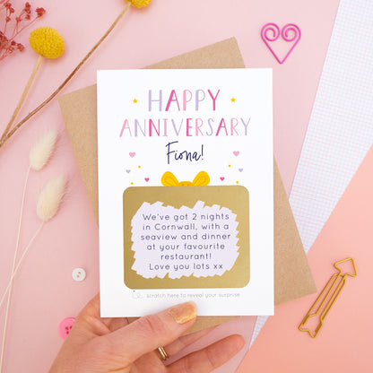 A personalised happy anniversary scratch card photographed on a pink background with floral props, paper clips, and buttons. The card has been scratched off to reveal the hidden message and is in the pink and lilac colour scheme. 