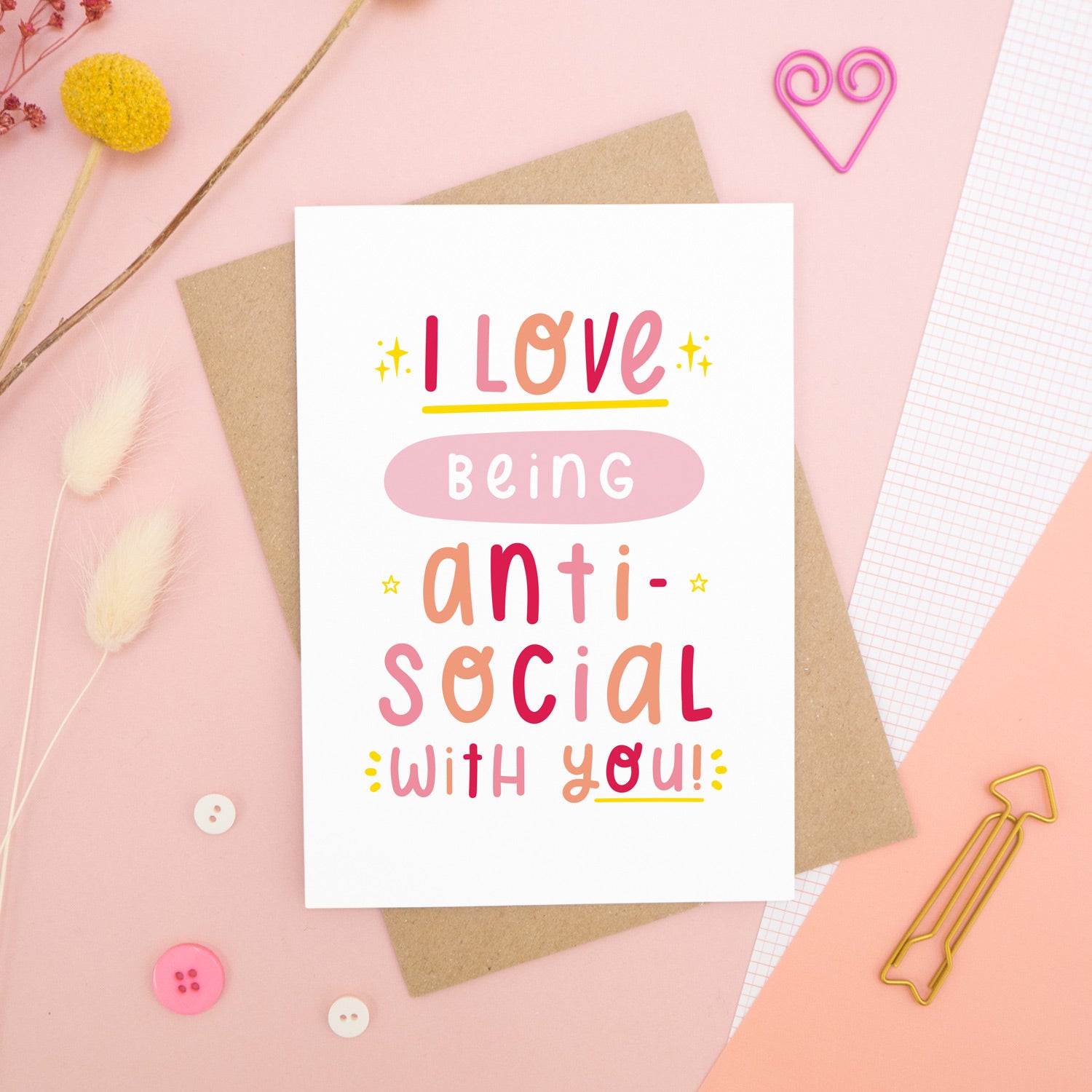The 'I love being anti-social with you' card photographed on a pink background with dried flowers, buttons and paper clips as props.