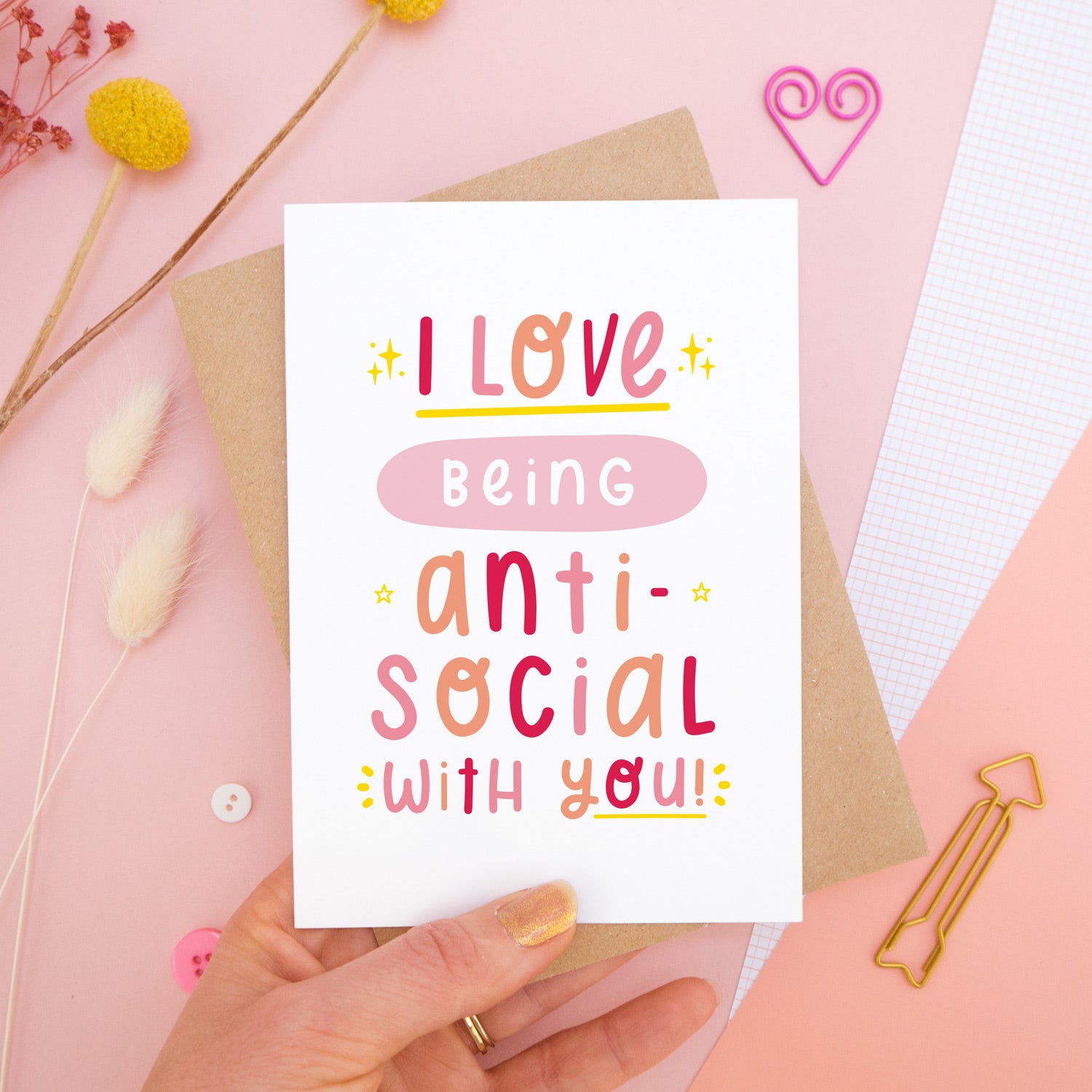 The 'I love being anti-social with you' card photographed on a pink background with dried flowers, buttons and paper clips as props. The card itself is being held above the scene.
