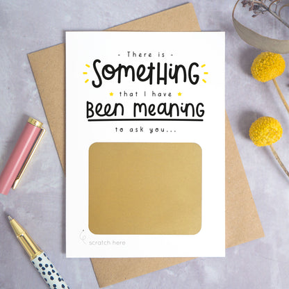 A personalised custom question scratch card photographed on a grey background with yellow flowers and a pink pen lid and spotty pen. The card is lying on a kraft brown envelope and the scratch box has not yet been scratched off. This is the black and yellow colour way.
