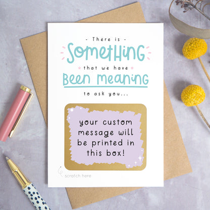 A personalised custom question scratch card photographed on a grey background with yellow flowers and a pink pen lid and spotty pen. The card is lying on a kraft brown envelope and the scratch box has been scratched off to reveal the personalised message. This is the teal and purple colour way.