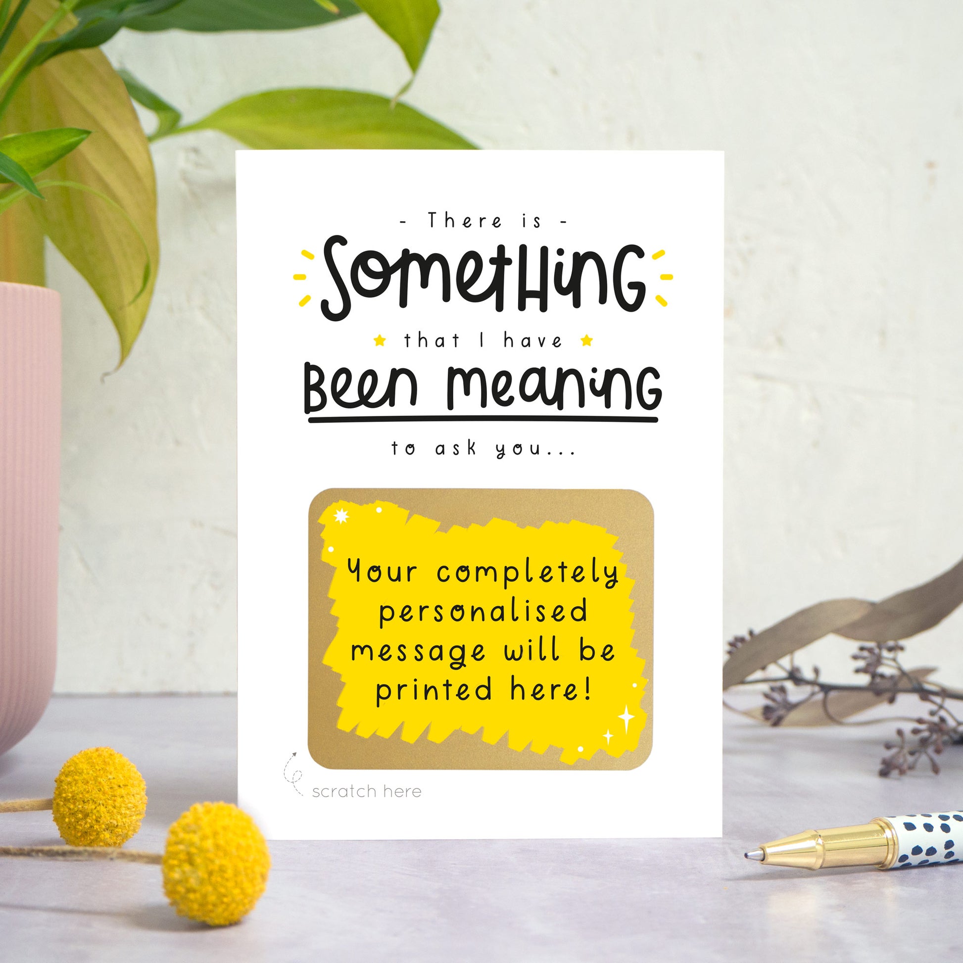 A personalised custom question scratch card photographed stood on a grey surface, a white textured background and with a pot plant on the left. Some small flowers and a pen are also in the foreground. This card is the black and yellow colour way with the scratch message revealed.
