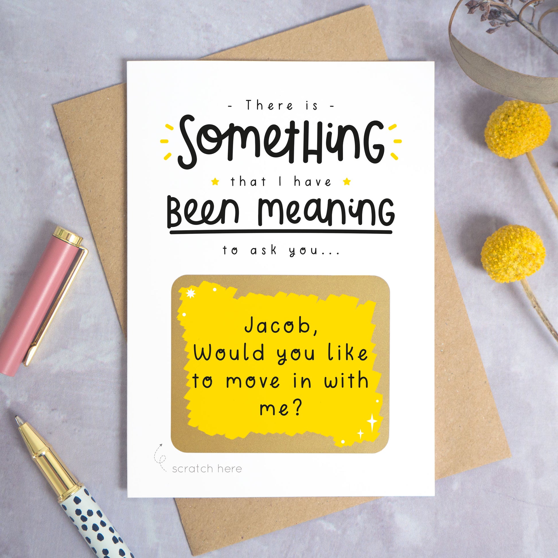 A personalised custom question scratch card photographed on a grey background with yellow flowers and a pink pen lid and spotty pen. The card is lying on a kraft brown envelope and the scratch box has been scratched off to reveal the personalised message. This is the yellow and black colour way.