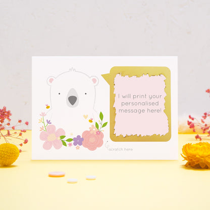 A personalised scratch card showing where your message will be printed featuring a white bear, pink flowers and a pink text box. This has been shot on a yellow and grey background with flowers. The gold panel has been scratched away to reveal the message.