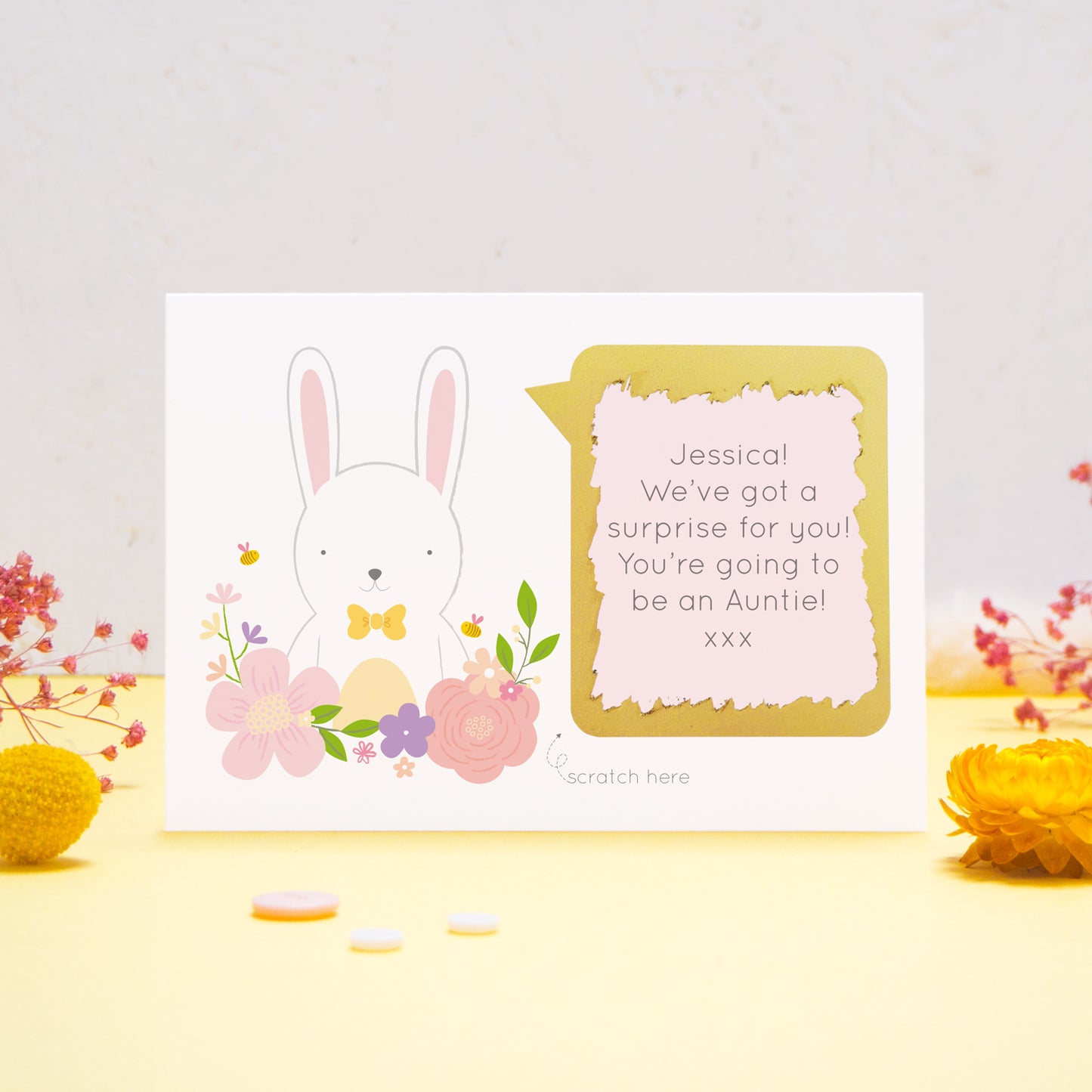 A personalised baby announcement scratch card featuring a white rabbit, pink flowers and a pink text box. This has been shot on a yellow and grey background with flowers. The gold panel has been scratched away to reveal the message.