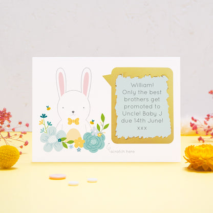 A personalised baby announcement scratch card featuring a white rabbit, blue flowers and a blue text box. This has been shot on a yellow and grey background with flowers. The gold panel has been scratched away to reveal the message.