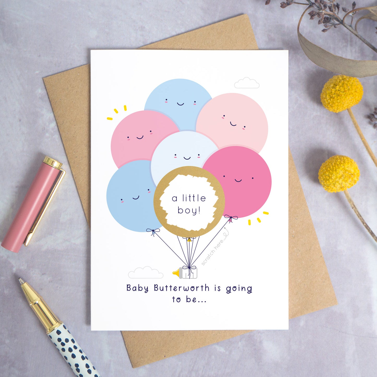 A ‘gender reveal’ scratch card photographed on a grey background with a pen on the left and yellow and purple flowers on the right. This is the pink & blue version of the card after it has been scratched off.