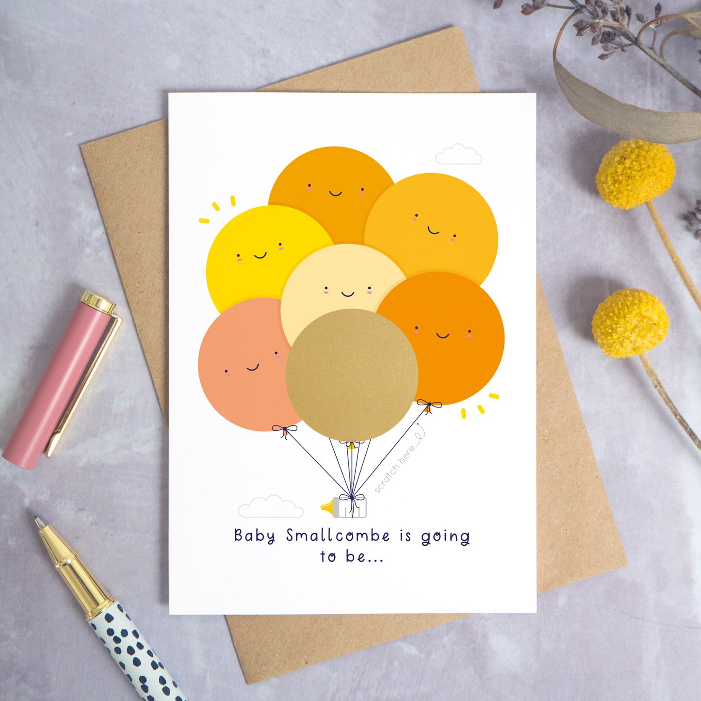 A ‘gender reveal’ scratch card photographed on a grey background with a pen on the left and yellow and purple flowers on the right. This is the yellow and orange version of the card before the gold panel has been scratched off.