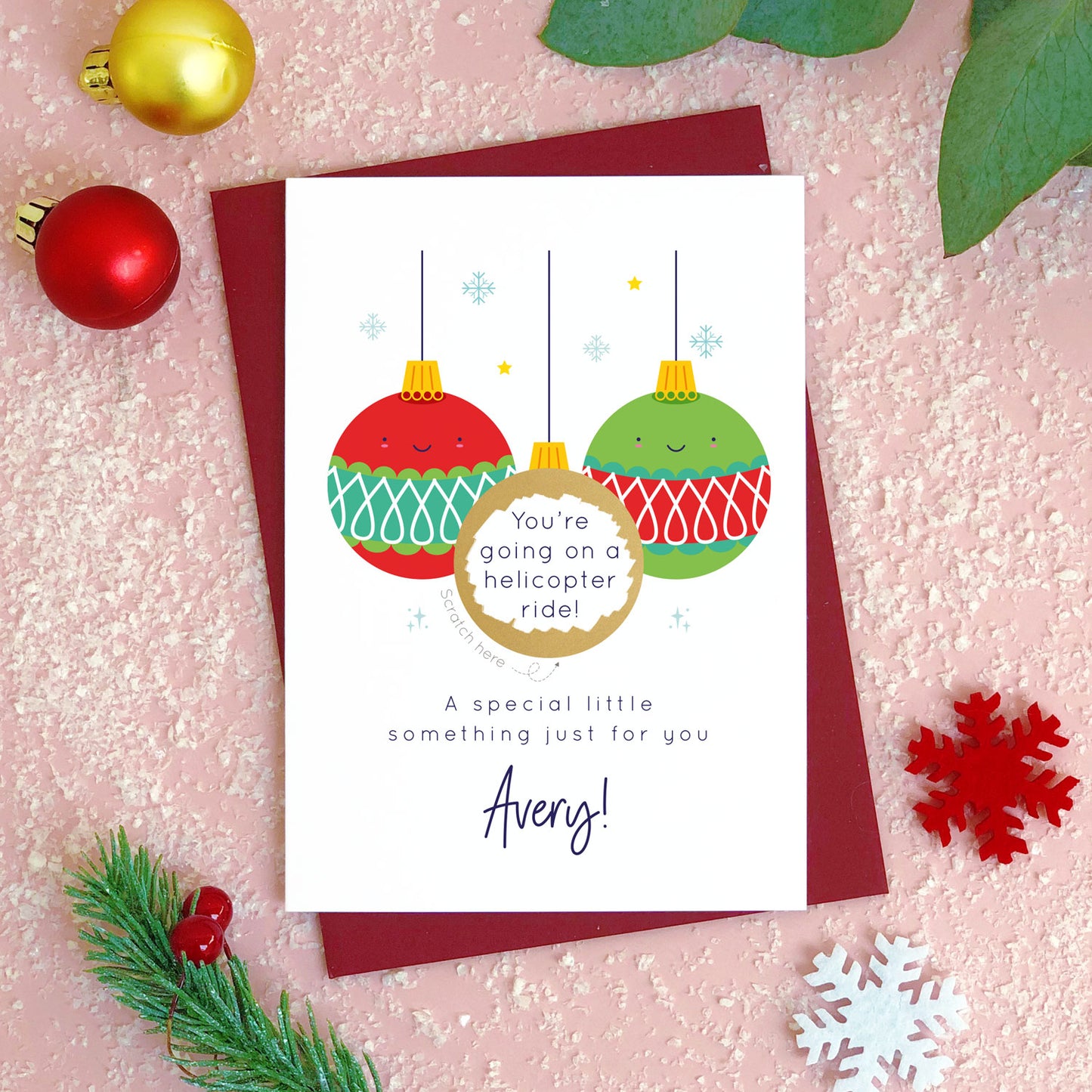 A personalised Christmas bauble scratch card photographed flat lying on a red wine coloured envelope on a pink surface surrounded by fake snow, baubles and foliage. The card is the green bauble version and the gold panel has been scratched off to reveal the custom message.