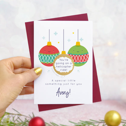 A personalised Christmas bauble scratch card photographed being held by a hand to the left and foliage and baubles in the bottom of the frame. The card is against its ‘red wine’ colour envelope and the design is the red and green version of the card. The scratch off panel has been scratched off to reveal the gift.