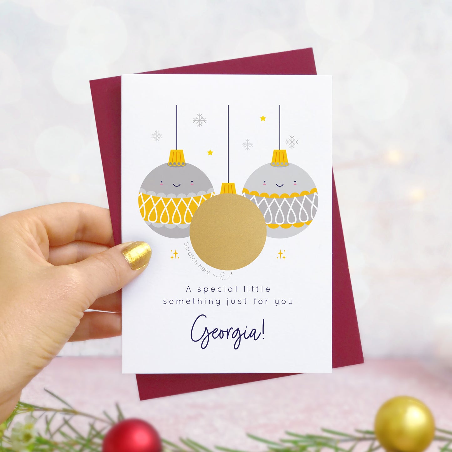 A personalised Christmas bauble scratch card photographed being held by a hand to the left and foliage and baubles in the bottom of the frame. The card is against its ‘red wine’ colour envelope and the design is the grey and yellow version of the card. The scratch off panel has yet to be scratched.