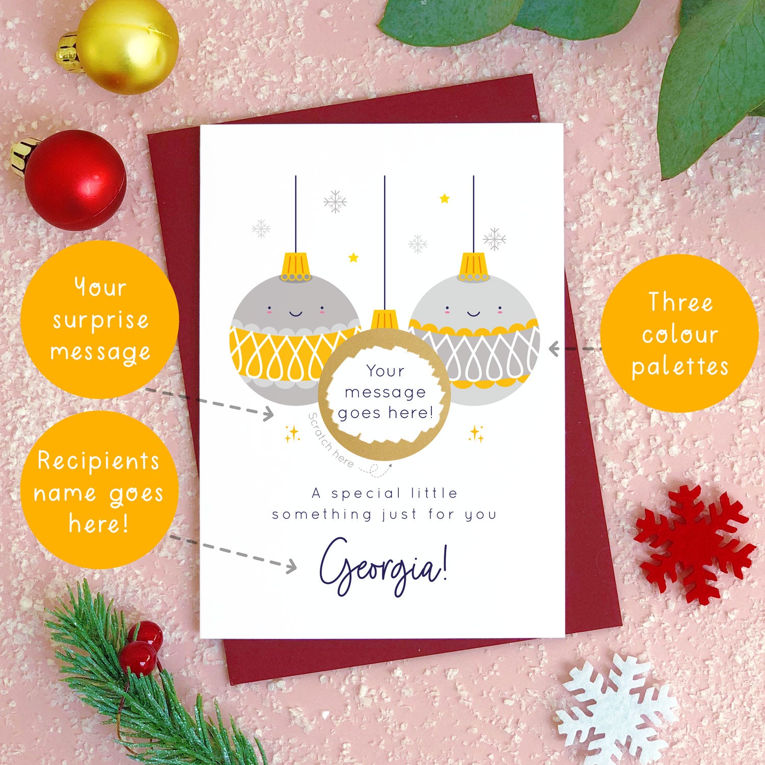 A personalised Christmas bauble scratch card photographed flat lying on a red wine coloured envelope on a pink surface surrounded by fake snow, baubles and foliage. The card is the grey and yellow bauble version. The orange circles demonstrate which areas of the card can be personalised. Here they point to the colour options, message and name for the front of the card.