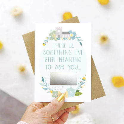 A will you be my godmother scratch and reveal card being held over a white background with pops of yellow. The design features a church, simple florals and a scratch off panel in silver. This is the blue palette.