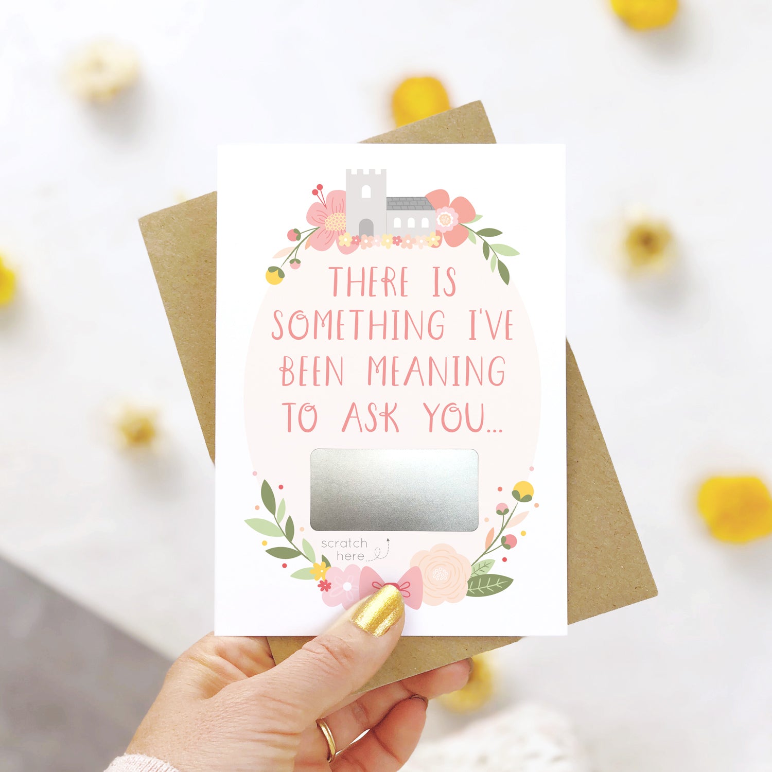 A will you be my godparents scratch and reveal card being held over a white background with pops of yellow. The design features a church, simple florals and a scratch off panel in silver. This is the pink palette.