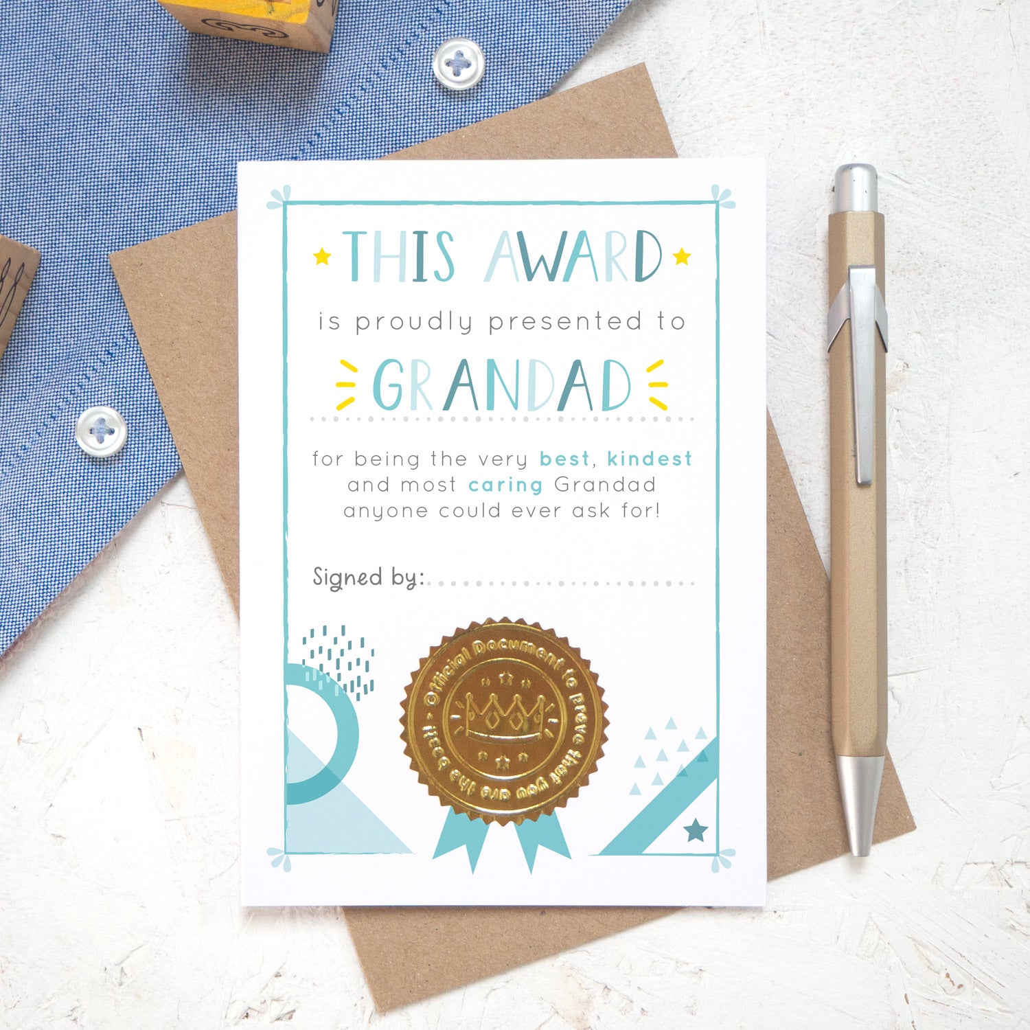 A Grandad certificate award card printed onto white card in varying tones of blue and pops of yellow! Each card features a shiny gold seal to make it official! Photographed on a white background with a hint of a blue shirt and a brown kraft envelope.